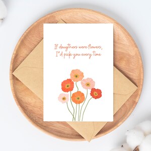 Peut inclure: Carte de v&oelig;ux avec le texte "Si les filles &eacute;taient des fleurs, je te choisirais &agrave; chaque fois" et une illustration florale orange et rose. La carte est pos&eacute;e sur un plateau en bois avec une enveloppe marron.