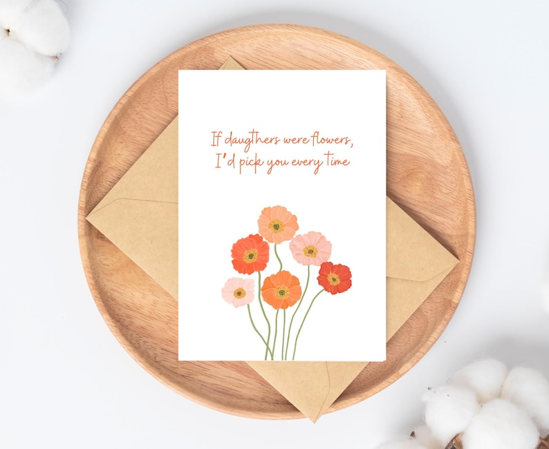 Peut inclure: Carte de v&oelig;ux avec le texte "Si les filles &eacute;taient des fleurs, je te choisirais &agrave; chaque fois" et une illustration florale orange et rose. La carte est pos&eacute;e sur un plateau en bois avec une enveloppe marron.
