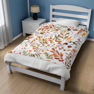 May include: A white twin bed with a floral patterned comforter. The comforter features a watercolor design of autumn leaves in shades of orange, green, and brown on a white background. The bed frame and headboard are white.