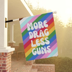 May include: A rainbow-colored flag with the text "More Drag Less Guns" in white, stylized lettering.
