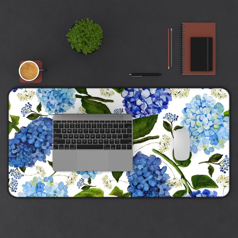 May include: A white desk mat with a blue and green floral pattern. The pattern features blue hydrangeas and green leaves on a white background. A laptop computer and a white mouse are on the desk mat.