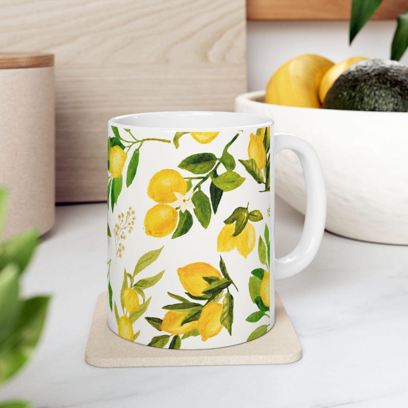 May include: White ceramic mug featuring a watercolor pattern of yellow lemons, green leaves, and white blossoms. The mug has a white handle and is placed on a beige coaster. The design is repeated around the entire mug.