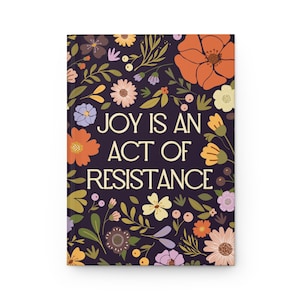 May include: A dark purple journal with a floral pattern and the text "JOY IS AN ACT OF RESISTANCE" in a light yellow font.