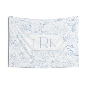 May include: A white tapestry with a blue floral pattern and the letters "LRK" in a rectangular frame. The tapestry features a delicate, botanical design with leaves and flowers.