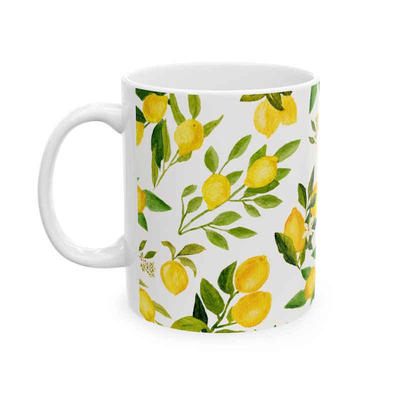 May include: White ceramic mug featuring a watercolor pattern of yellow lemons and green leaves on a white background. The mug has a curved handle and a cylindrical shape, suitable for hot or cold beverages.