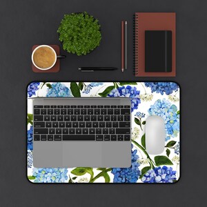May include: A white desk mat with a blue and green floral pattern featuring hydrangeas. A laptop computer, a mouse, and a cup of coffee are on the mat.