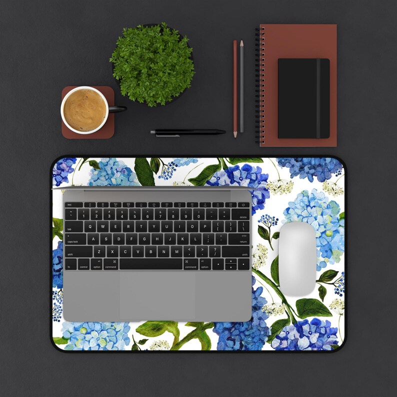 May include: A white desk mat with a blue and green floral pattern featuring hydrangeas. A laptop computer, a mouse, and a cup of coffee are on the mat.