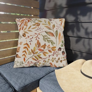 May include: A throw pillow with a cream-colored background and a watercolor pattern of fall leaves in shades of orange, yellow, and green. The pillow is sitting on a blue couch.
