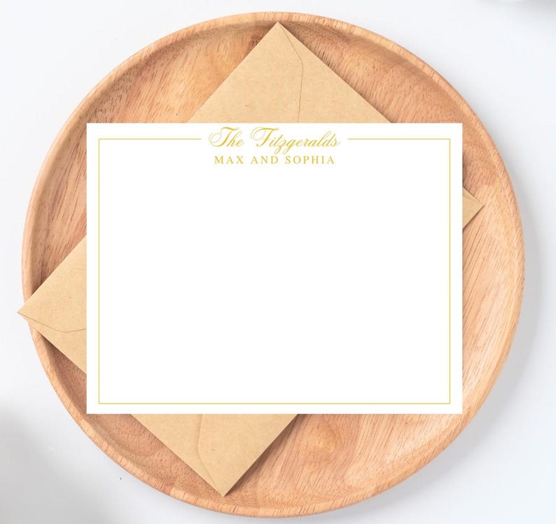 May include: A white stationery card with a gold border and the text "The Fitzgeralds MAX AND SOPHIA" is displayed on a wooden plate with a brown envelope. The stationery is blank, ready for personalization.