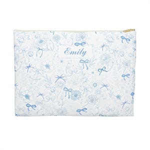 May include: A white zipped pouch featuring a light blue floral and bow design. The pouch has a gold zip and the name "Emily" printed in light blue on a white rectangle.