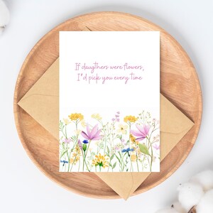 Peut inclure: Une carte de v&oelig;ux avec un motif floral et le texte "If daughters were flowers, I'd pick you every time." La carte est sur un plateau en bois avec une enveloppe en papier kraft.