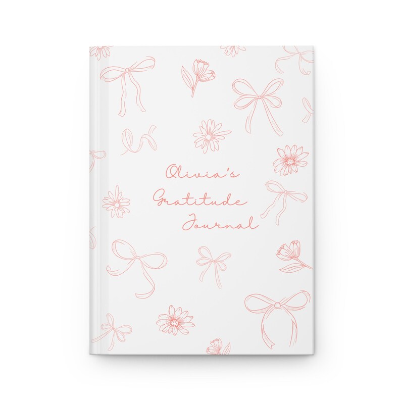 May include: A gratitude journal with a white cover features a blush pink design of bows and daisies. The text on the cover reads 'Olivia's Gratitude Journal' in a script font. This personalized journal is perfect for expressing thankfulness and practicing gratitude.