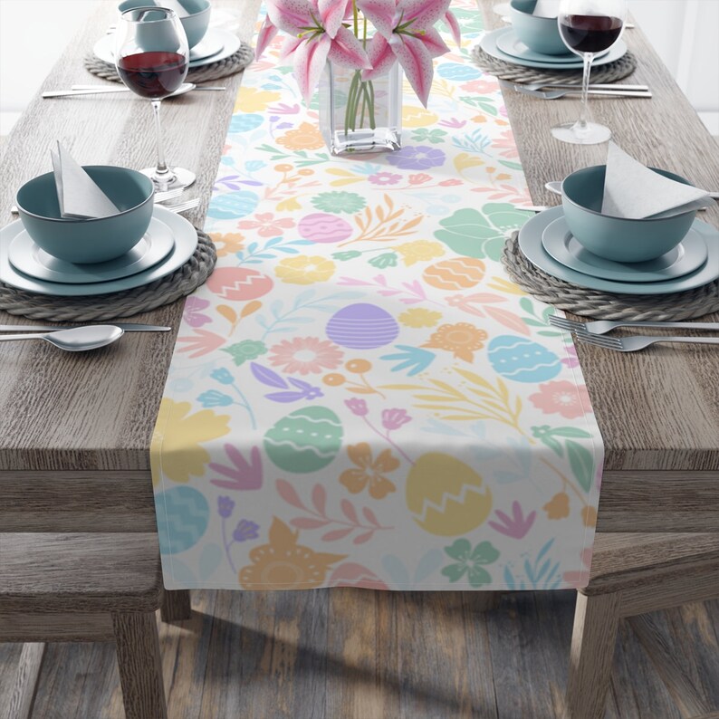 May include: A white Easter-themed table runner with a pastel floral and egg pattern. The runner is placed on a wooden table set for a meal, with plates, glasses, and silverware visible.