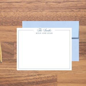 May include: A stationery set featuring a white card with a blue border and the text "The Smiths, Kyle and Leah." A light blue envelope is behind the card. A gold pen is on the wooden surface.
