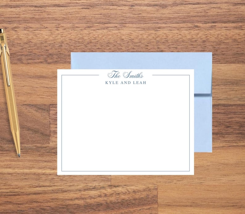 May include: A stationery set featuring a white card with a blue border and the text "The Smiths, Kyle and Leah." A light blue envelope is behind the card. A gold pen is on the wooden surface.