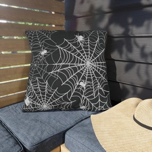May include: Black square throw pillow with a white spiderweb design and several white spiders. The pillow is on a blue cushion, with a wooden slatted background and a straw hat in the foreground. Halloween decor.