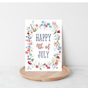 May include: A white card with a wreath of red, blue, and pink flowers surrounding the text "HAPPY 4th of JULY" in blue and red lettering.