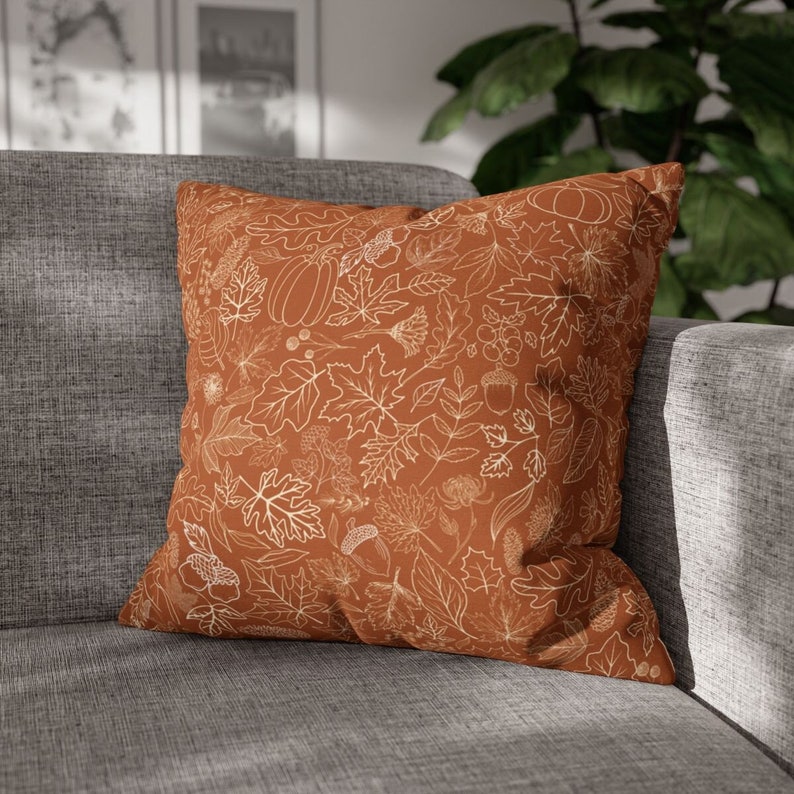 May include: A brown throw pillow with a white outline of fall leaves, pumpkins, and acorns. The pillow is on a gray couch.