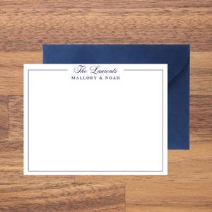 May include: A white stationery card with a navy blue border and the text "The Laurents" above "Mallory & Noah" in a script font. A navy blue envelope is behind the card, set against a wooden surface.