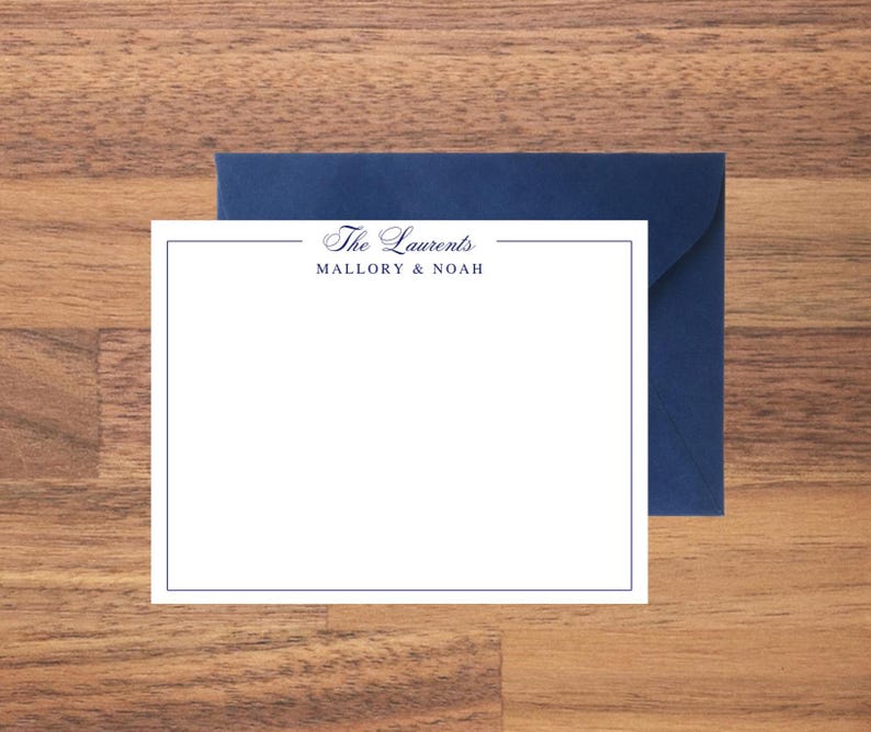 May include: A white stationery card with a navy blue border and the text "The Laurents" above "Mallory & Noah" in a script font. A navy blue envelope is behind the card, set against a wooden surface.