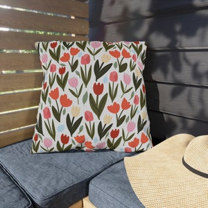 May include: A white pillow with a floral pattern featuring red, pink, and yellow tulips on a white background. The pillow is sitting on a blue couch with a straw hat.