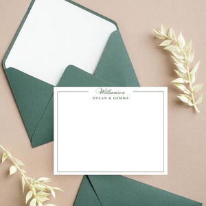 May include: A stationery set featuring a white card with a dark green border and the names "Williamson, Dylan & Gemma" printed in dark green. The card is surrounded by dark green envelopes and decorative sprigs on a tan surface.