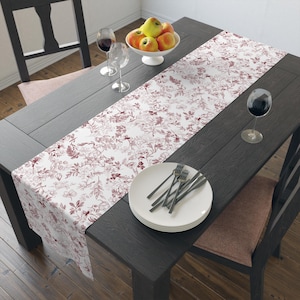 May include: A dining table set with a floral patterned table runner, white plates, and silverware. The table is set with wine glasses and a bowl of apples. The table and chairs are dark wood.
