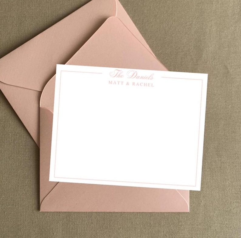 May include: A blush pink stationery set featuring a blank card with a double border and matching envelope. The card is printed with "The Daniels" and "MATT & RACHEL" in a delicate script. The set is displayed on a neutral background.
