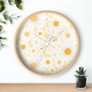 May include: A round wall clock with a light wood frame. The clock face features a pattern of white daisies with yellow centers on a pale yellow background. White clock hands indicate the time. The clock is on a wooden surface.