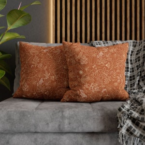 May include: Two brown throw pillows with a white outline of fall leaves, pumpkins, and acorns. The pillows are on a gray couch with a black and white throw blanket.