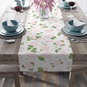 Puede incluir: Un camino de mesa blanco con un estampado floral de flores rosas y hojas verdes. El camino de mesa está colocado sobre una mesa de madera, con cubiertos y un jarrón con flores rosas. La mesa está puesta para una comida.