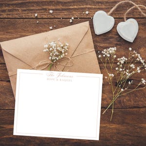 May include: A stationery set featuring a blank white card with a gold border, an envelope, and a sprig of baby's breath. The text "The Johnsons HOPE & RAQUEL" is printed at the top of the card. Two heart-shaped ornaments hang from a twine string.