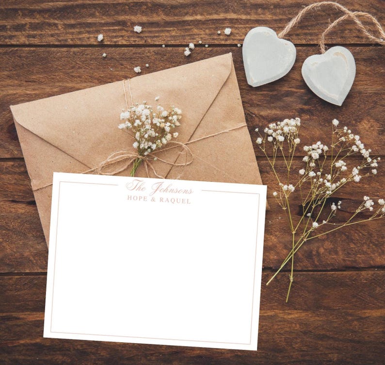 May include: A stationery set featuring a blank white card with a gold border, an envelope, and a sprig of baby's breath. The text "The Johnsons HOPE & RAQUEL" is printed at the top of the card. Two heart-shaped ornaments hang from a twine string.