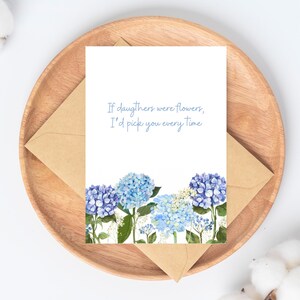 Peut inclure: Carte de v&oelig;ux avec des fleurs d'hortensia bleues et le texte "If daughters were flowers, I'd pick you every time." La carte est pos&eacute;e sur une assiette en bois avec une enveloppe marron.