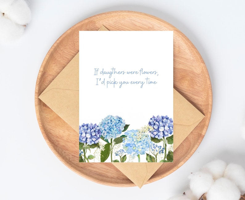 Peut inclure: Carte de v&oelig;ux avec des fleurs d'hortensia bleues et le texte "If daughters were flowers, I'd pick you every time." La carte est pos&eacute;e sur une assiette en bois avec une enveloppe marron.