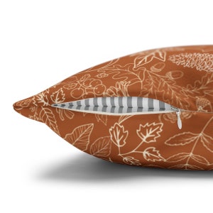 May include: A rust-colored throw pillow with a white outline of leaves, berries, and acorns. The pillow has a zipper closure and a white and gray striped lining.