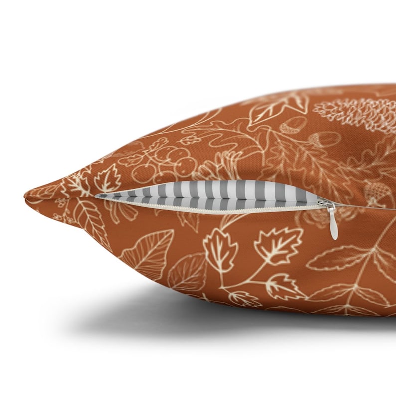 May include: A rust-colored throw pillow with a white outline of leaves, berries, and acorns. The pillow has a zipper closure and a white and gray striped lining.