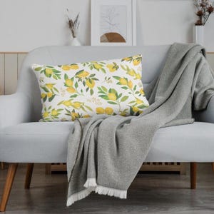 May include: A decorative rectangular pillow with a lemon and leaf pattern rests on a light gray loveseat. A gray blanket with fringe is draped over the loveseat. The pillow features yellow lemons and green leaves.