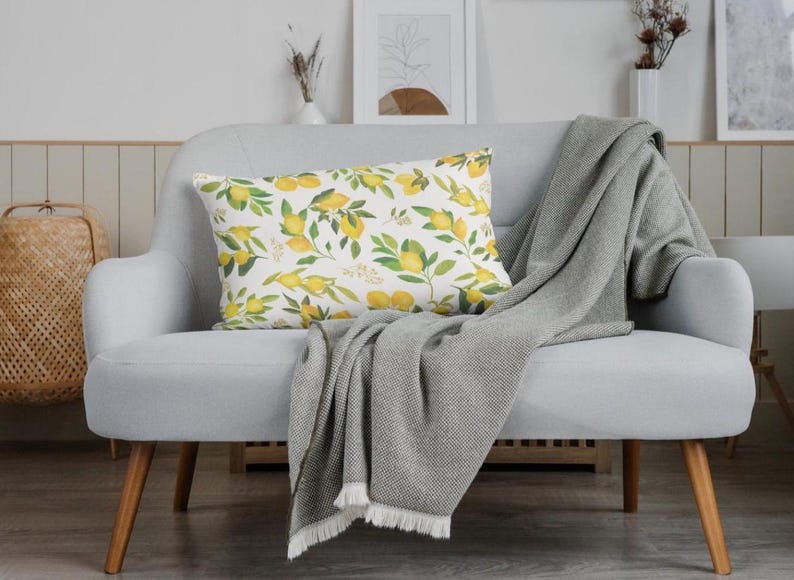 May include: A decorative rectangular pillow with a lemon and leaf pattern rests on a light gray loveseat. A gray blanket with fringe is draped over the loveseat. The pillow features yellow lemons and green leaves.