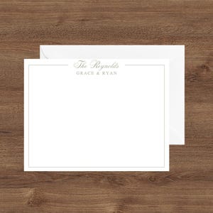May include: White stationery set with a matching envelope. The stationery has a thin gold border and the text "The Reynolds" and "Grace & Ryan" in a delicate font. The set is displayed on a wooden surface.