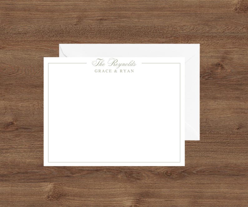 May include: White stationery set with a matching envelope. The stationery has a thin gold border and the text "The Reynolds" and "Grace & Ryan" in a delicate font. The set is displayed on a wooden surface.
