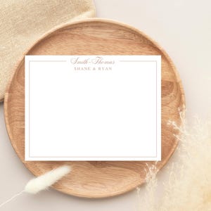 May include: A white stationery card with a gold border and text, "Smith-Thomas" and "SHANE & RYAN", rests on a wooden plate. A beige linen cloth and dried ornamental grass are in the background.