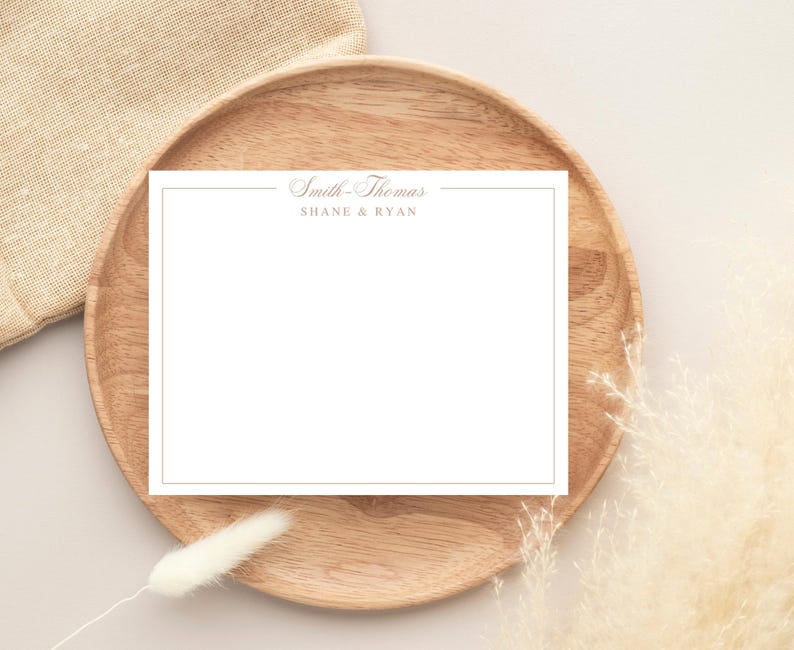 May include: A white stationery card with a gold border and text, "Smith-Thomas" and "SHANE & RYAN", rests on a wooden plate. A beige linen cloth and dried ornamental grass are in the background.