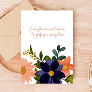 Peut inclure: Carte de v&oelig;ux blanche avec le texte "Si les filles &eacute;taient des fleurs, je te choisirais &agrave; chaque fois" et des illustrations florales roses, oranges et bleues. Une enveloppe en papier kraft est pos&eacute;e sur un plateau en bois.