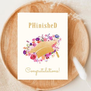 May include: A white card with the text "PHinished" in gold lettering. A gold graduation cap is surrounded by pink, purple, and orange watercolor flowers. The text "Congratulations!" is written in gold lettering below the flowers.