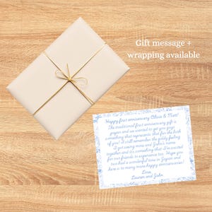 May include: A wrapped gift with a cream-colored paper and gold string tied in a bow. A card with blue floral accents and handwritten text is next to the gift. The text on the card reads "Happy first anniversary Olivia & Matt!"