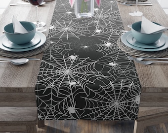 May include: A black table runner featuring a white spiderweb design with spiders. The runner is placed on a wooden table set for a meal, with plates, bowls, and glasses visible. Pink flowers in a vase add a pop of color.