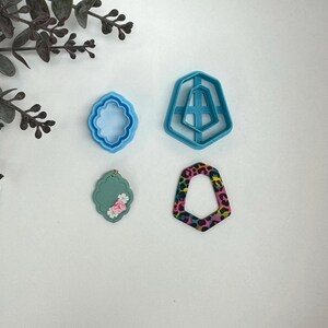 May include: A set of four cookie cutters, including a geometric shape, a floral design, a rectangle with a floral design, and a leopard print design. The cutters are blue and pink.