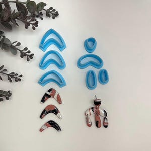 May include: A set of blue polymer clay cutters in the shape of arches and ovals. The cutters are arranged in two groups. The first group has three arches of different sizes. The second group has one oval, one arch, and two smaller ovals. Below the cutters are three polymer clay earrings in the shape of arches, painted in shades of brown, black, and white. The earrings are arranged in a similar pattern to the cutters.