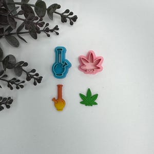 May include: Four colorful cookie cutters in the shapes of a bong and a cannabis leaf. The bong cutters are blue and yellow, and the leaf cutters are pink and green. The cutters are on a white surface.